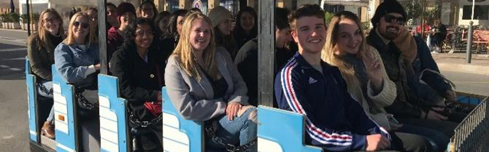 Students are riding in a small tram car through the streets of Aix. They are looking at the camera and smiling.