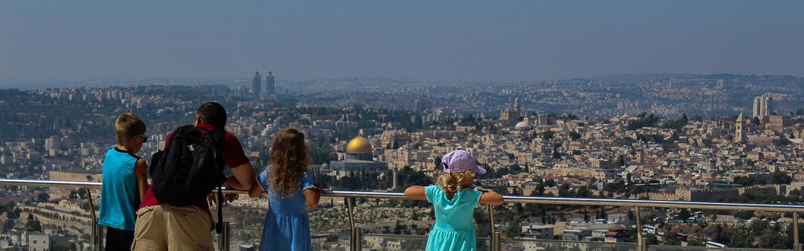 A man and three kids overlook a balcony to see a city with many tall buildings, including one central building with a golden dome. 