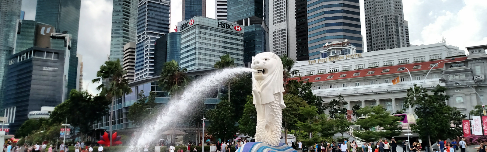 A white fountain in the shape of a Merlion, a create with the head of a lion and body of a mermaid. Tall glass buildings are behind it.