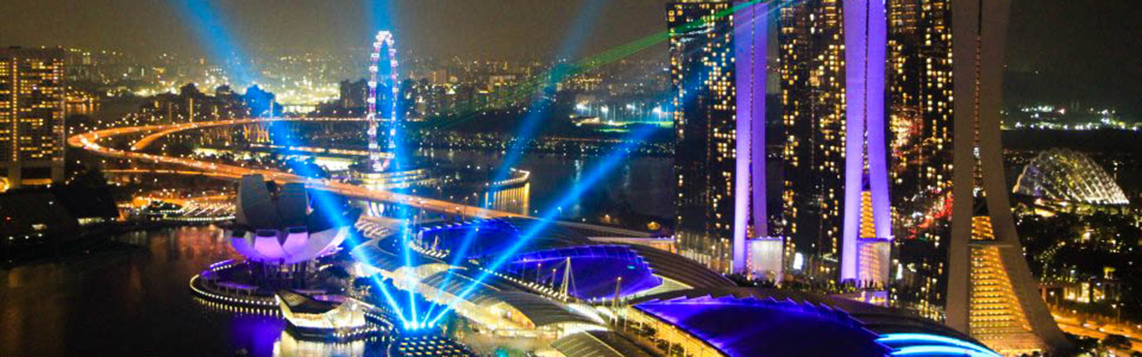 An aerial view of Singapore at night, with a light show and the Marina Bay Sands towers in the foreground.