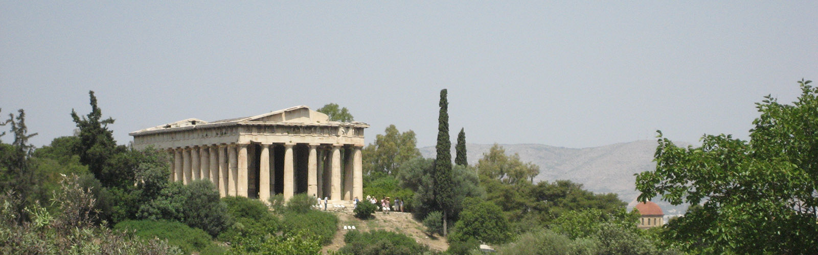 Hiltop view of an ancient Greek temple. The temple is surrounded by trees and other greenery, and some people are standing outside the temple.