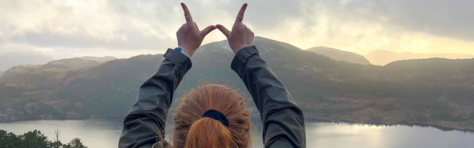 A person stands in front of a lake with the "finger W" pose, surrounded by mountains that are glowing in the sunset.