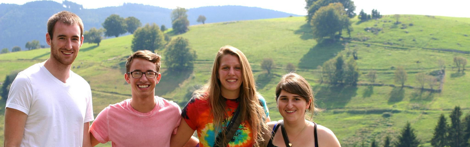 Four students are at the base of a hill. There are trees and other greenery in the background. They are all smiling at the camera.