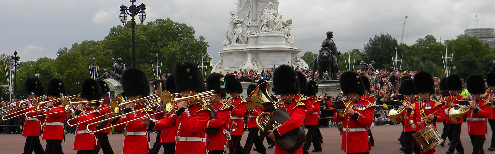 Marching band of British soldiers in their iconic red uniforms marching around the streets near Buckingham Palace. There are crowds gathered on the sides.