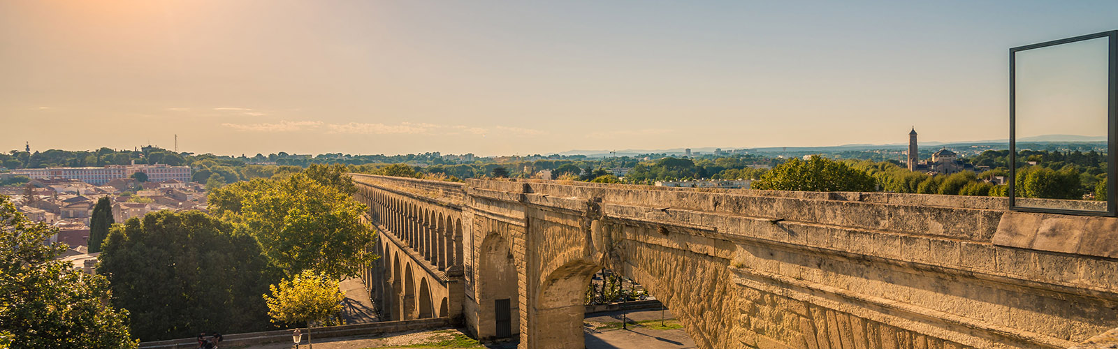 Long bridge through Montpellier. It has old arches for support and is surrounded by greenery. The city is in the background.