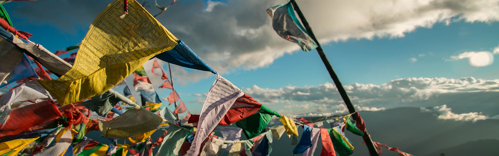 Countless vibrant flags are waving in the wind near the mountains. They are somewhat worn down.