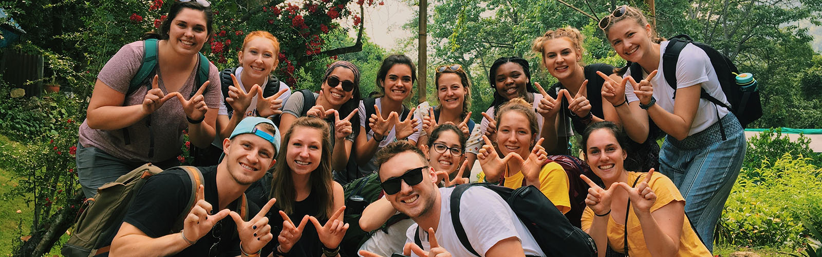 Twenty students stand in a wooded area and throw up the "finger W" pose. 