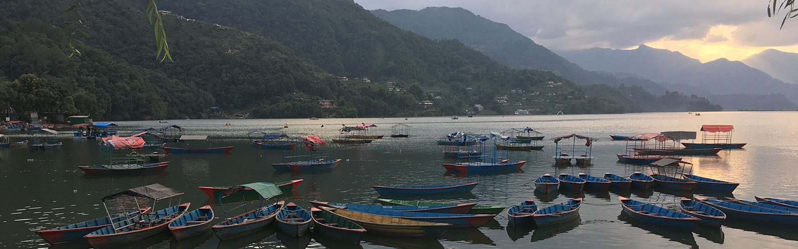 Many small boats, mostly blue canoes, sit on a lake as the sun peeks out from behind the mountains in the distance. 