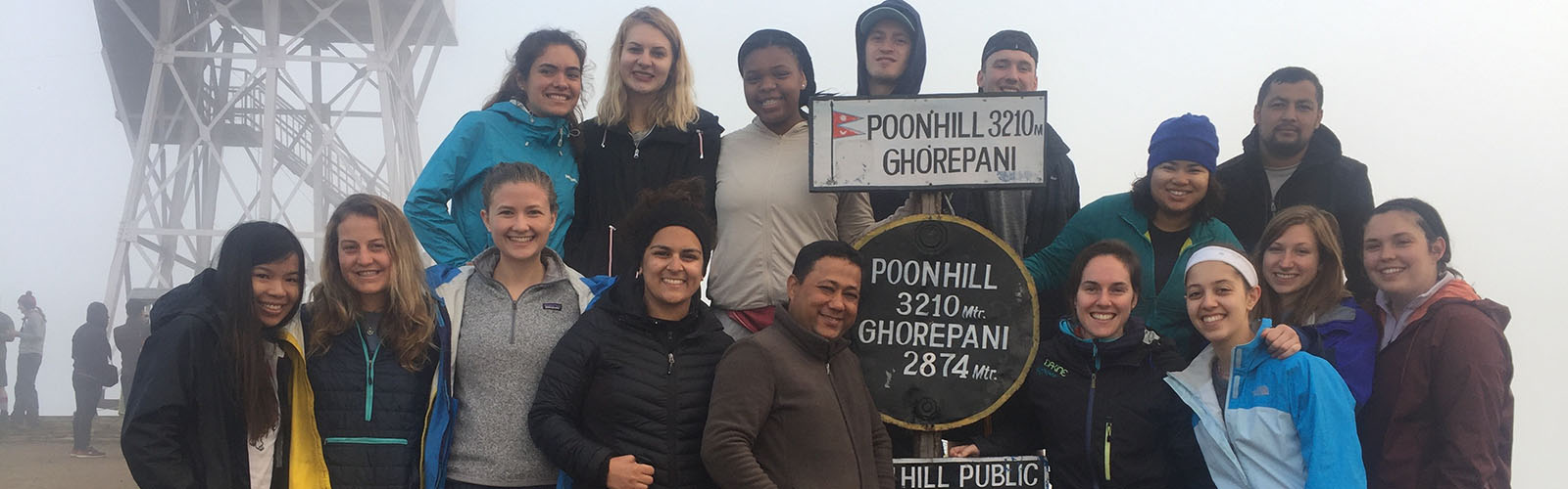 A group of students in warm clothes stand around a sign that says "Poon Hill". 
