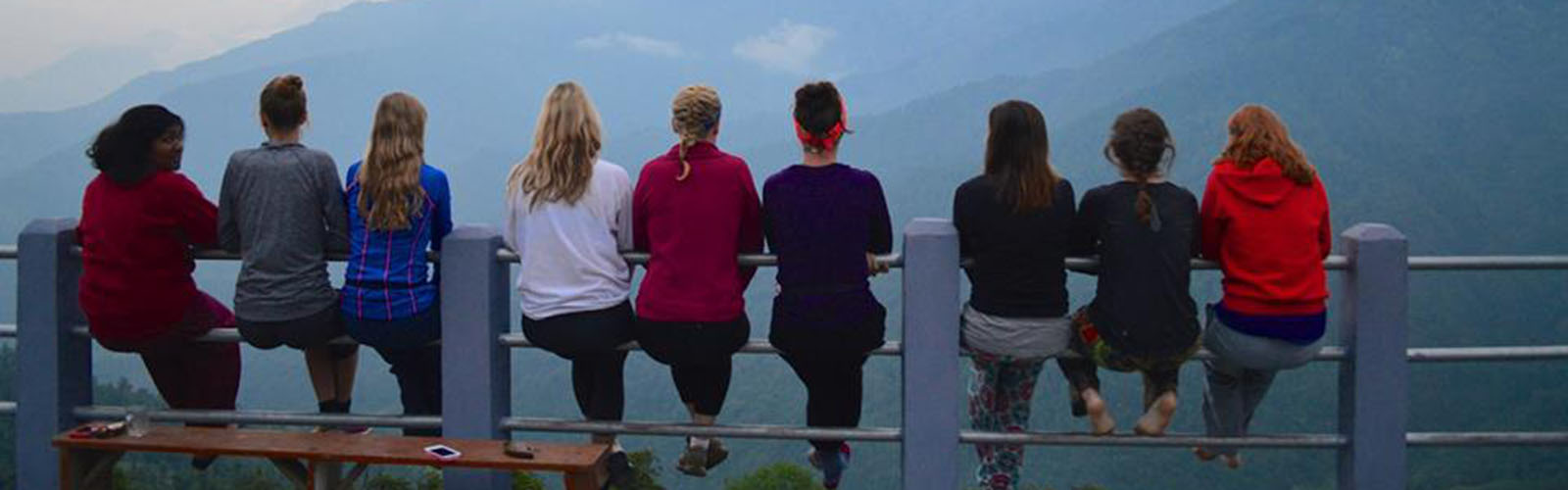 A group of students as seen from the back sitting on a metal railing overlooking many mountains. 