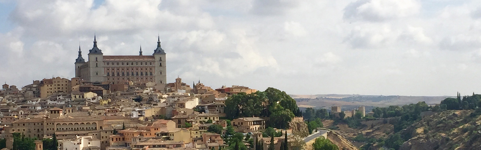 Tan and beige buildings packed densely on a hillside. A large, rectangular building with pointed towers on the corners sits at the top.