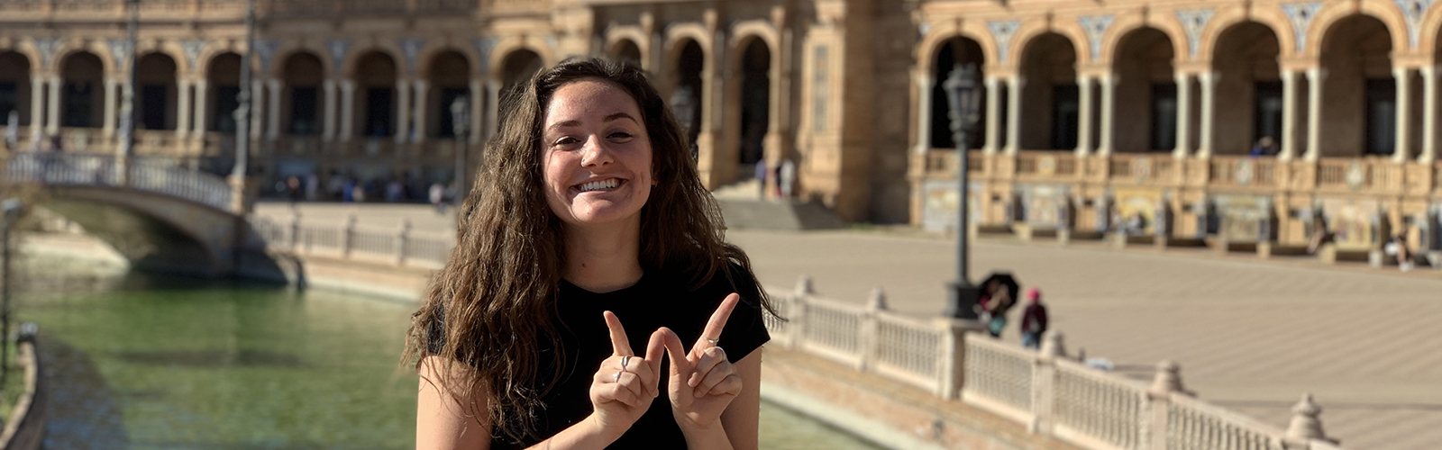 A student, standing on a bridge over a small waterway, holding up the letter "W" with their fingers. A curving, columned building is a distance behind them.