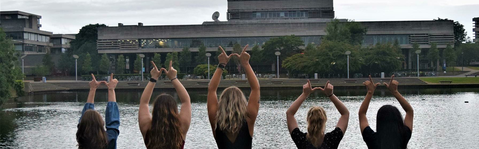 Five students, photographed from the back, are standing in front of a river doing the "Finger W" pose.