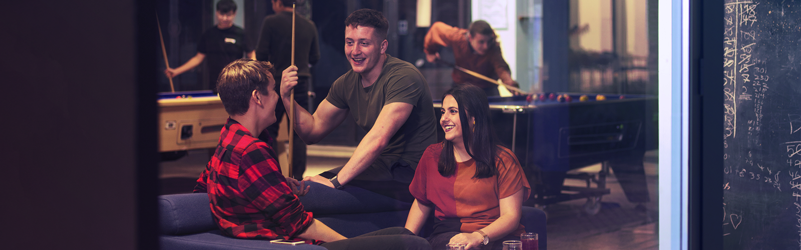 Three students are hanging out and playing pool. They are looking at eachother and smiling.