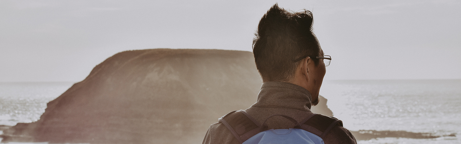 A student is looking away from the camera and out into the ocean. A large rock is in their view.