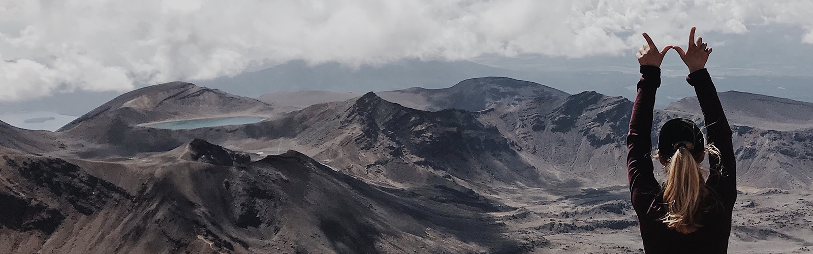 A student stands facing rolling mountains, holding up the "finger W" high above her head. 