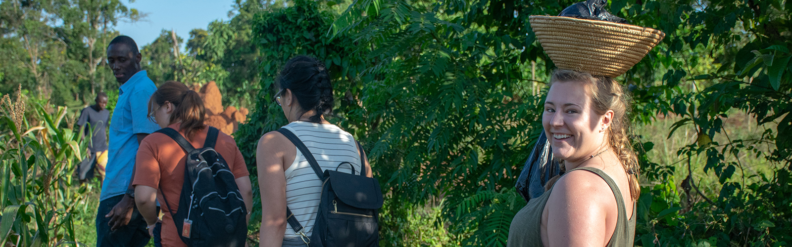 Three students led through a farm by a guide. One student is balancing a basket on their head and is smiling at the camera.