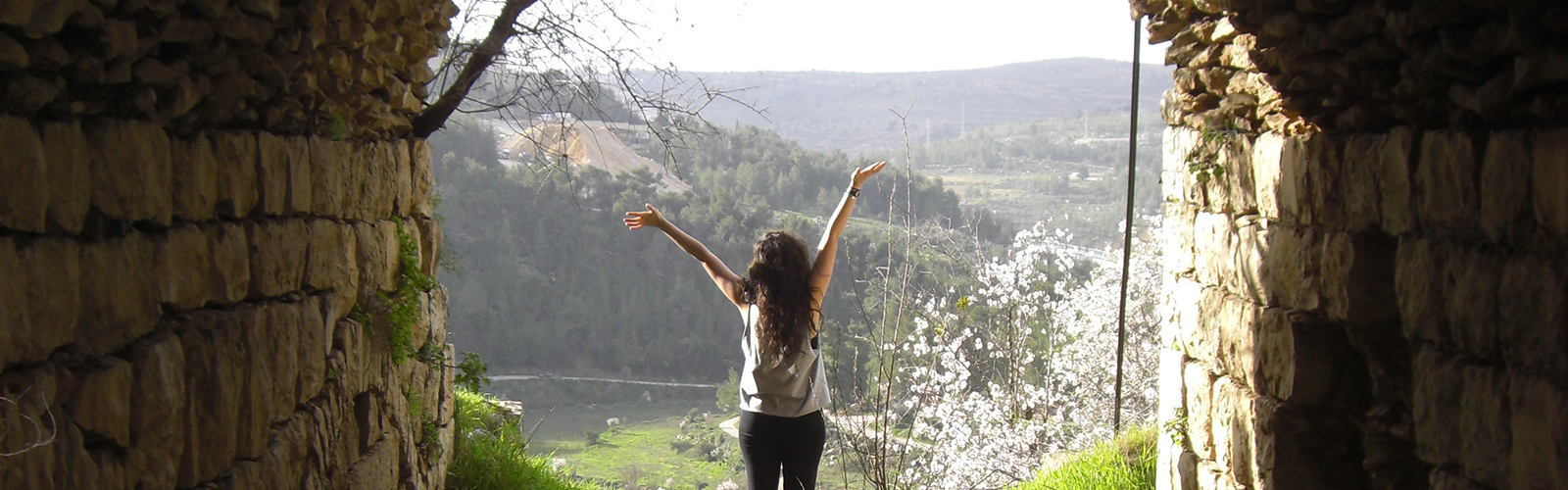 An opening of an old, stone building, where a person stands with their arms raised overlooking a green landscape with mountains, a river, and flowering trees. 