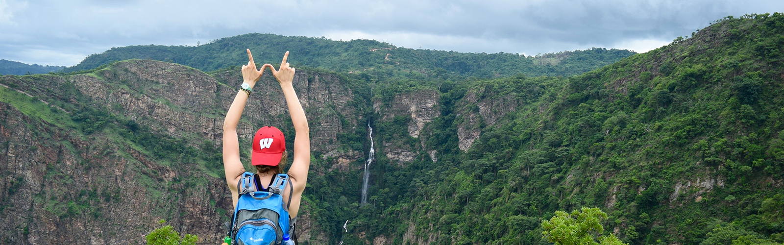 A student is on top of a mountain and overlooking a vast forest. They are making a W sign with their hands and facing away from the camera.