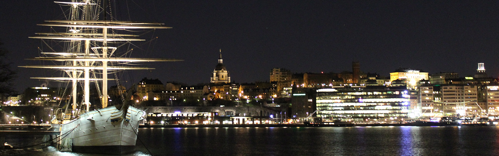 A white boat with many masts floating in a harbor at night. Brightly lit modern buildings take up the background.