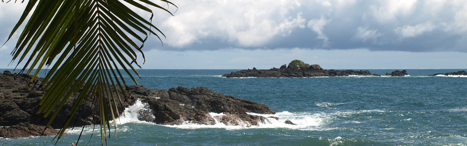 Waves are crashing onto small Islands. A palm leaf is sticking out in the foreground.