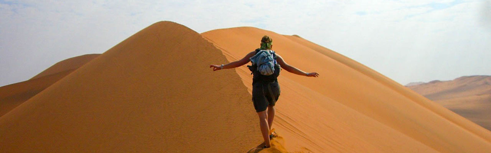 An individual walking along the ridge of a large sand dune. More dunes fill the background.