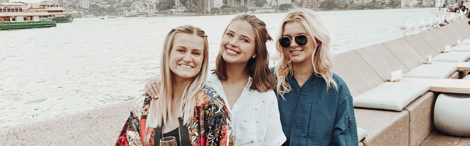 Three students are standing outside near the water. They are all smiling at the camera.