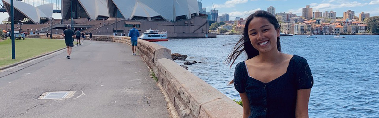 A student is in front of the Sydney Opera House and is smiling at the camera. Behind them is the Sydney skyline and the water.