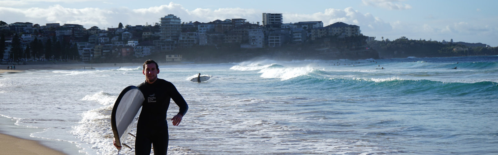 A surfer is walking out of the ocean. Heavy waves are crashing in the background.