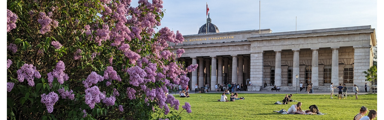 Large old building with many pillars. People are on the lawn of the building, and a bush with flowers indicates that it is springtime.