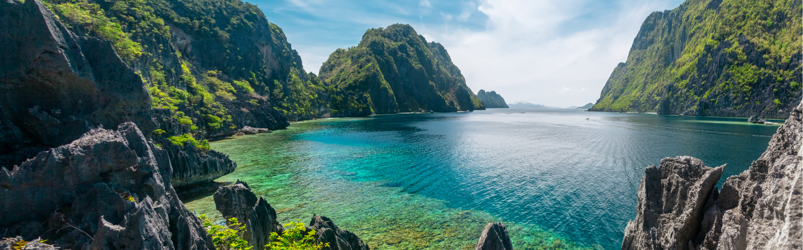 Turquoise coastal inlet framed by rugged, green-covered cliffs, with clear water, rocky foreground, and distant mountains under a bright sky.
