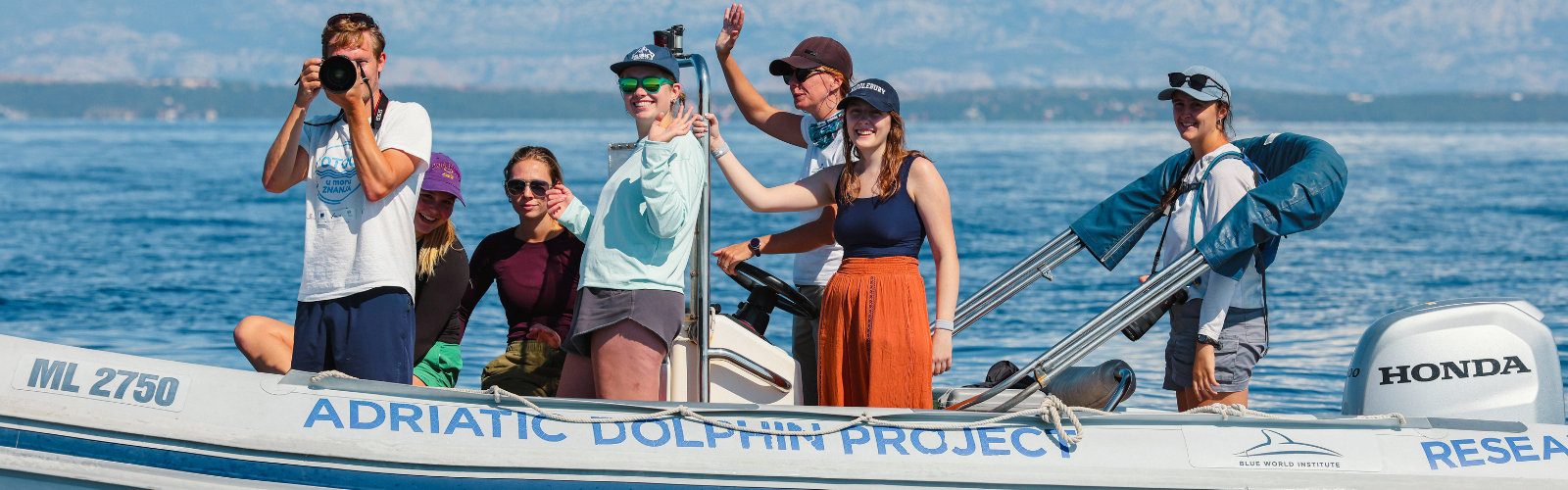 Students are on a small boat. One is taking photos and others are waving at the camera. 