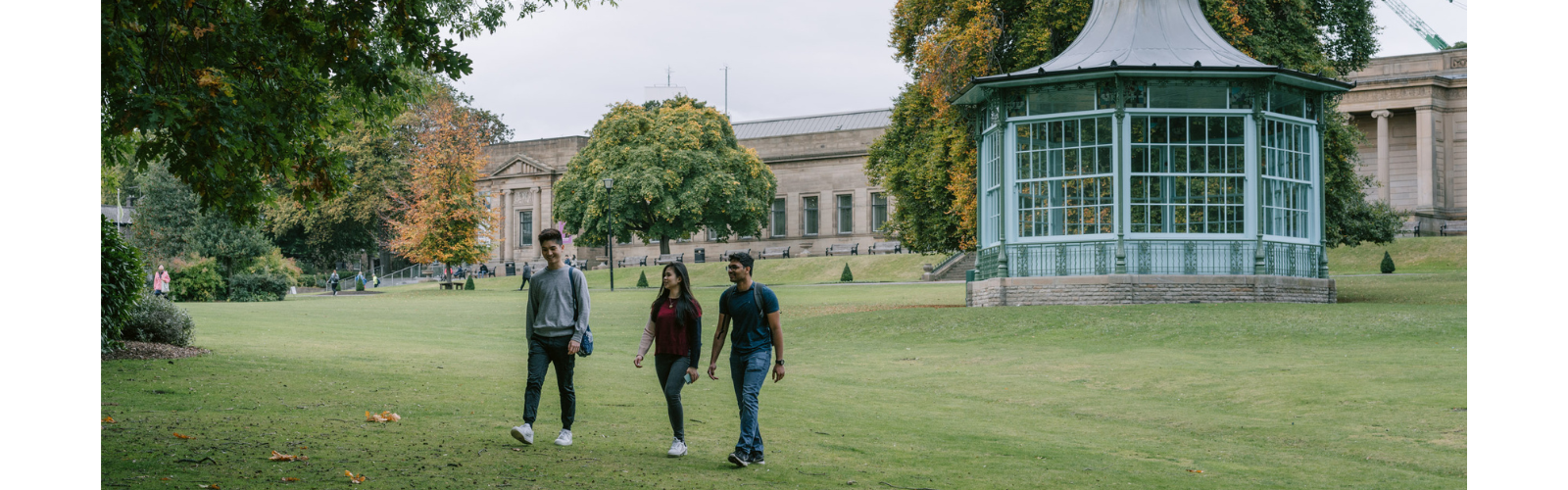 Three students are walking on the lawn of the campus. The university buildings are in the background and the students are looking at each other.