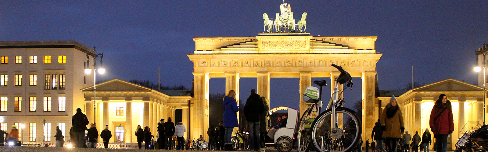Iconic archway in the center of Berlin is surrounded by people. It is nighttime.