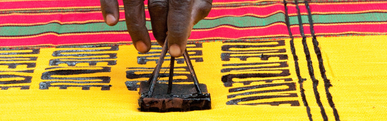 Zoomed in view of an artist stamping dark letters onto a piece of colorful fabric. Only their hand is visible.