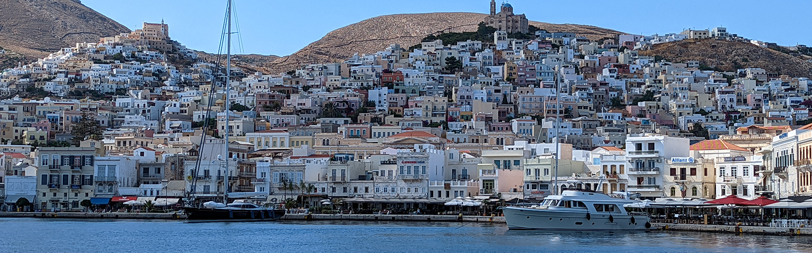 View of a city just off the coast of the island. There are boats in the foreground and buildings escalading down the hill on land.