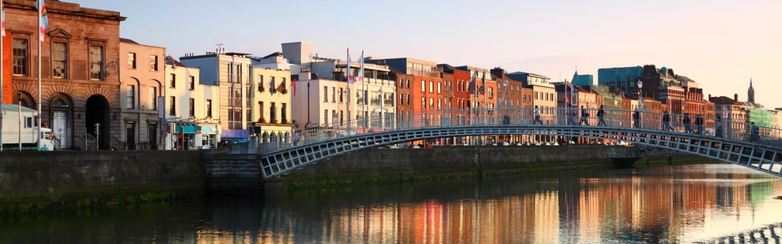 Several colorful buildings along the edge of a river are dimly lit by the rising sun. A metal bridge crosses the river. 
