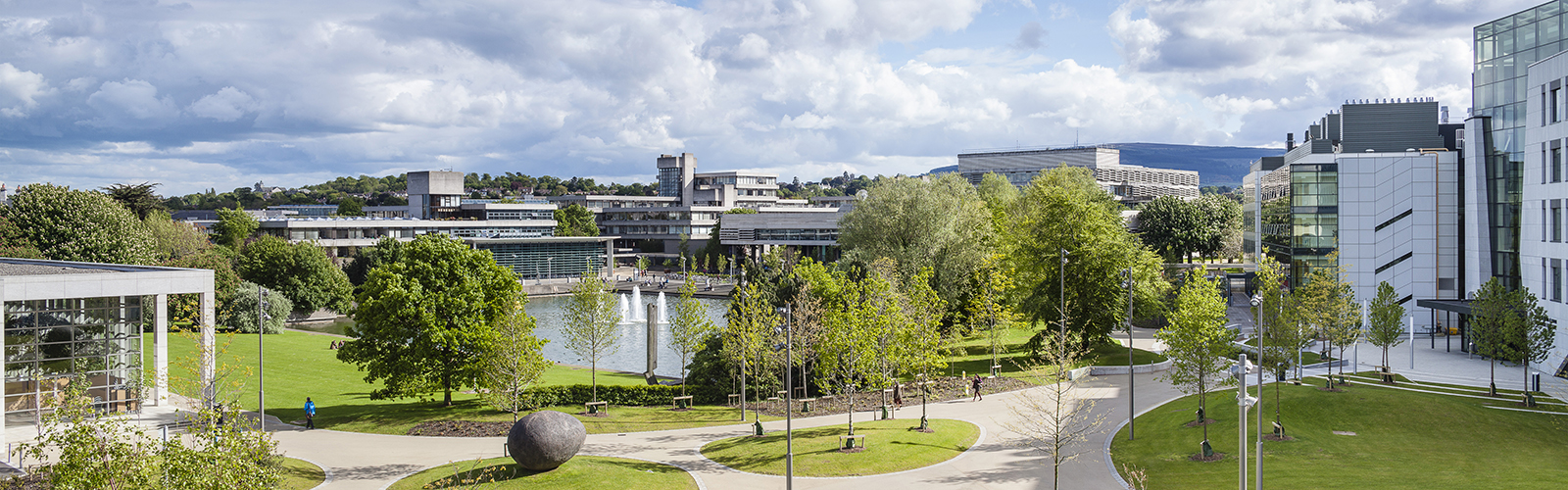 Many modern on-campus buildings, surrounded by patches of green grass, trees, and a pond. 