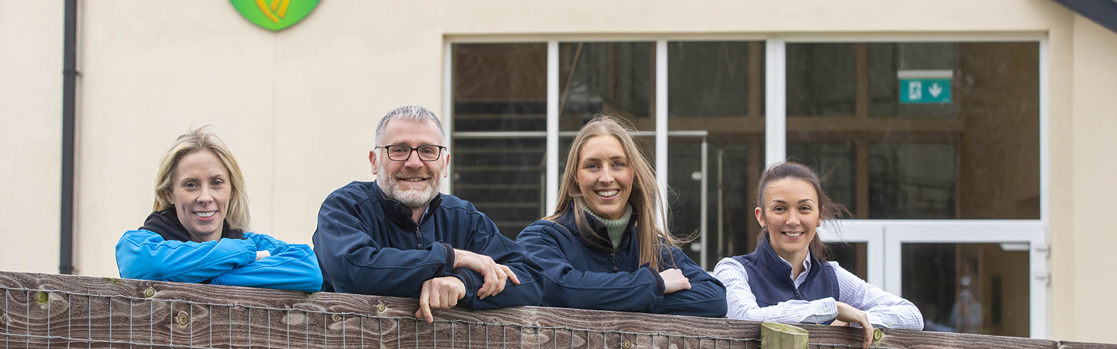 Four people are leaning over a wooden fence with a building in the background.