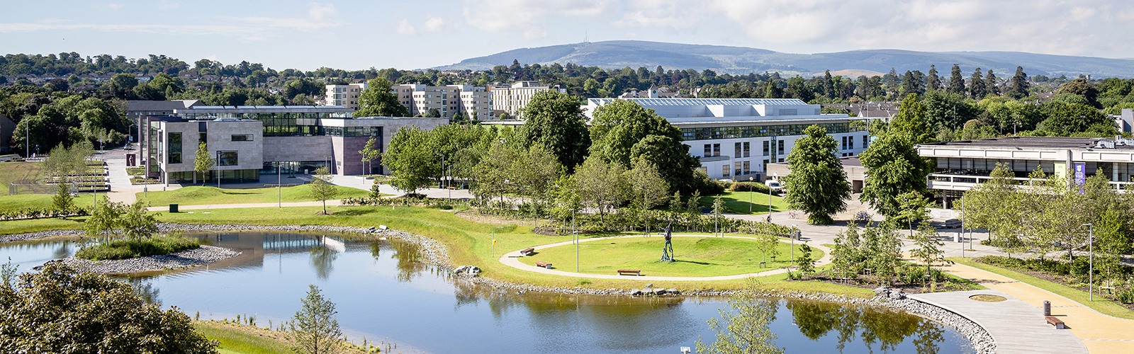 A modern building with a glass roof sits behind a body of water, and there are trees and mountains behind it. 