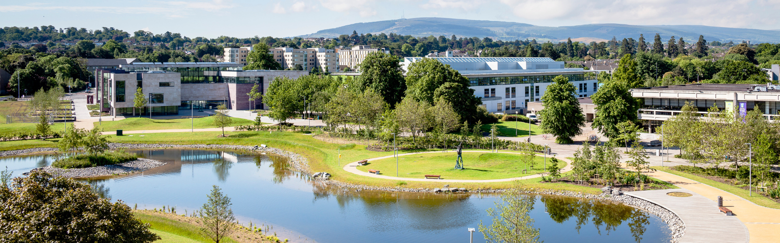 A modern building with a glass roof sits behind a body of water, and there are trees and mountains behind it. 