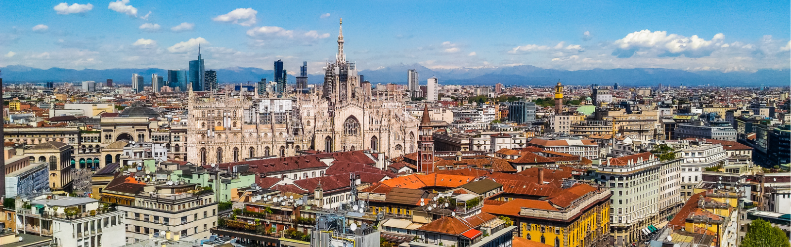 Aerial view of Milan's skyline featuring the Duomo Cathedral with its intricate spires. Surrounding buildings display varied architecture under a clear blue sky.