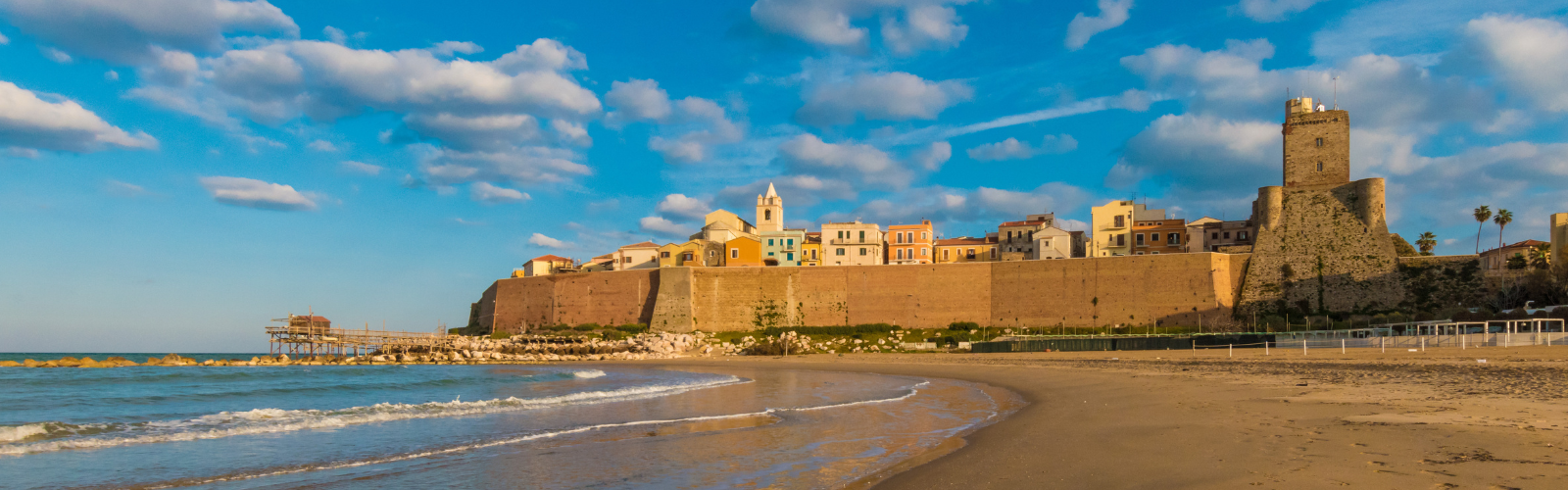A sandy beach meets water in the foreground, with a large stone wall separating the water from a city full of colorful buildings in the background. 