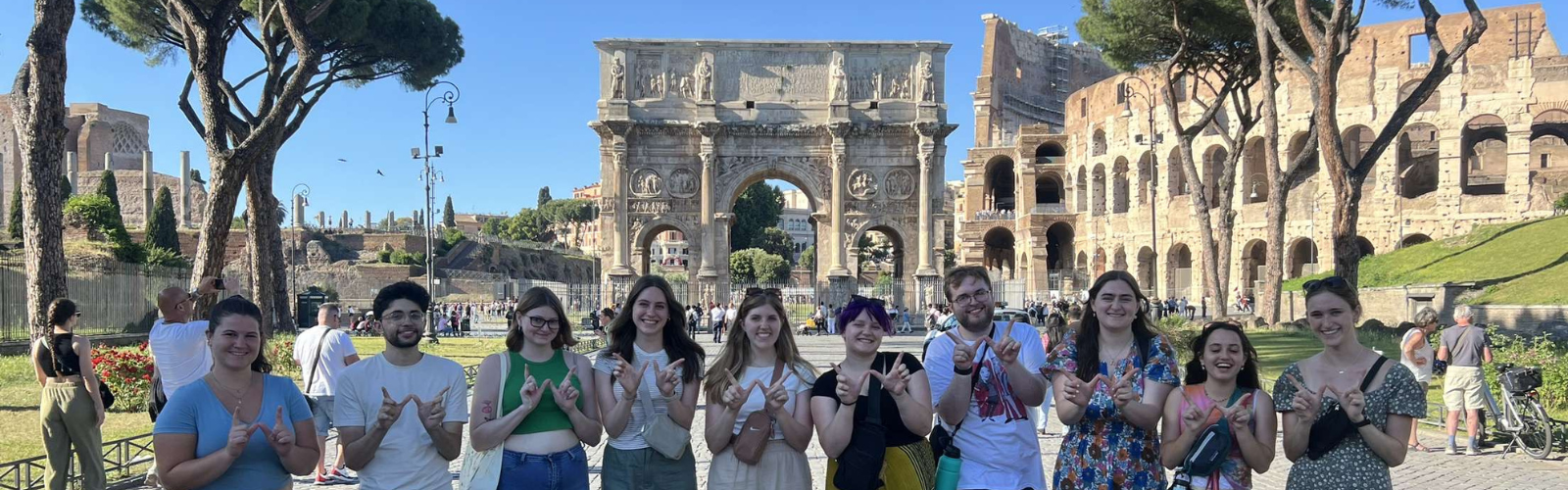 A line of ten students doing the "finger W" pose in front of multiple stone ruins with many archways. 