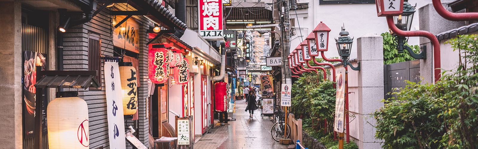 A narrow street with many stores packed closely together with black, red, and yellow exteriors on the left side, and many red street lights on the right side. 