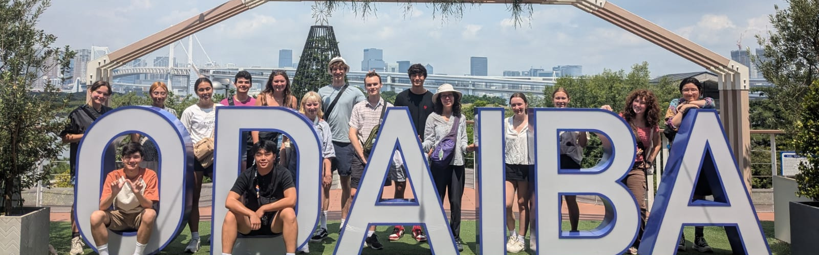 A group of several students stand around a blue and white sign that says "Odabia", with a student sitting inside both the letter "O" and the letter "D". They are standing in front of a cityscape. 
