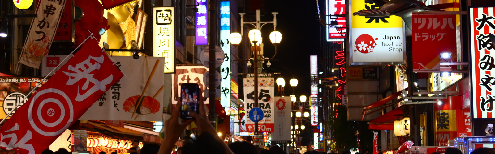 An urban street in Japan at night. Many signs in kanji illuminate a dense crowd below them.