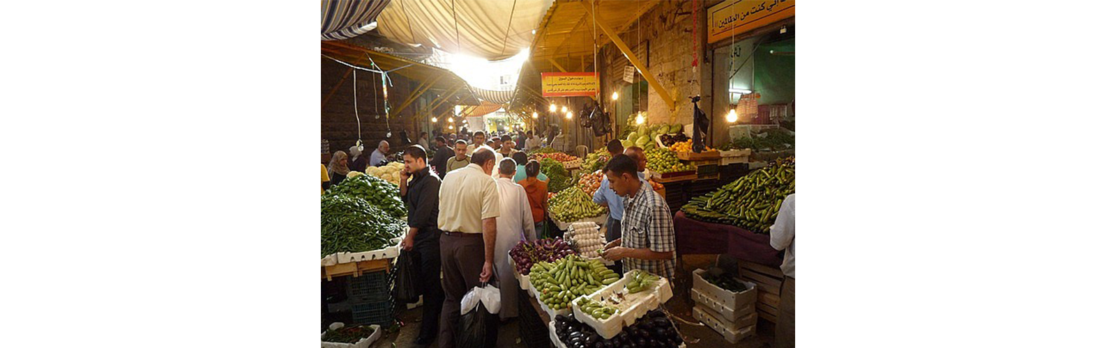 A marketplace in an enclosed area that is packed with citizens and vendors. Wooden crates are piled high with different green vegetables. 