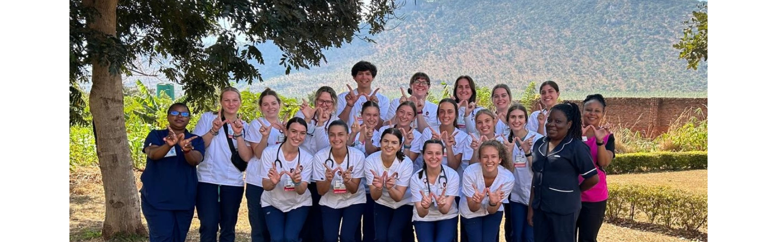 Three rows of students wearing stethoscopes are posing for the camera and showing the "finger W" in front of bushes and a mountain. 