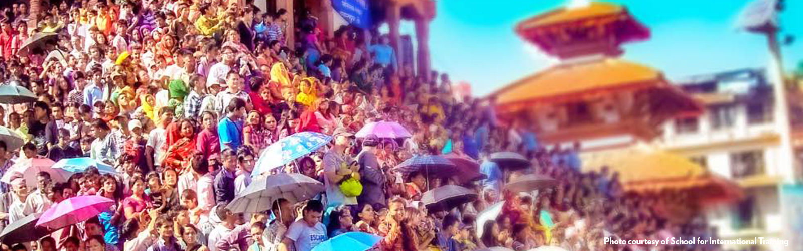 A large crowd of people in colorful clothing standing in front of a building with tiered roofs. Some people are holding colorful umbrellas. 